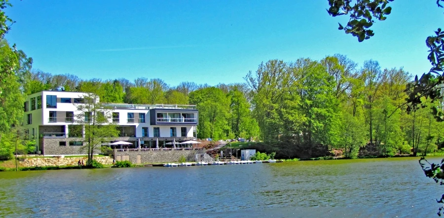 Ein modernes weißes Gebäude mit Balkonen blickt auf einen ruhigen See, der an einem sonnigen Tag von üppigen grünen Bäumen umgeben ist - der perfekte Ort für einen Last-Minute-Urlaub, mit Tischen und Stühlen am Wasser und kleinen Booten, die am Ufer angedockt sind.