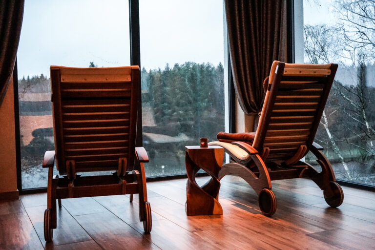 Zwei Liegestühle aus Holz und ein kleiner Tisch stehen vor großen Fenstern mit Blick auf eine regnerische Waldlandschaft und schaffen so eine gemütliche und friedliche Atmosphäre im Innenraum im Peters Hotel & Spa