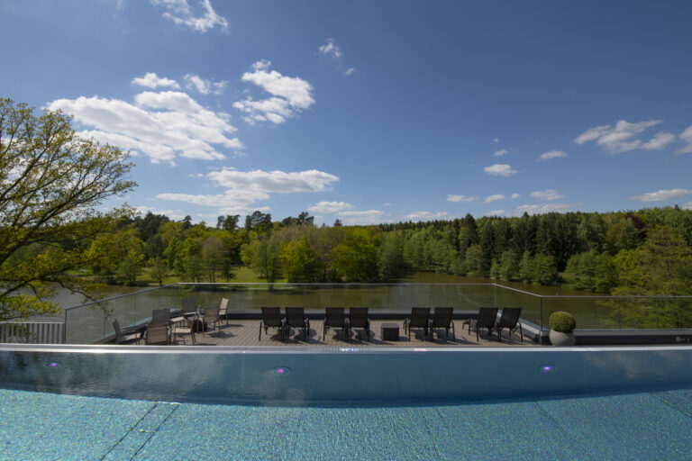 Ein Infinity-Pool mit kristallklarem blauem Wasser blickt auf eine Sonnenterrasse mit Liegestühlen, umgeben von üppig grünen Bäumen und einem ruhigen See unter einem strahlend blauen Himmel mit vereinzelten Wolken. Im Peters Hotel & Spa