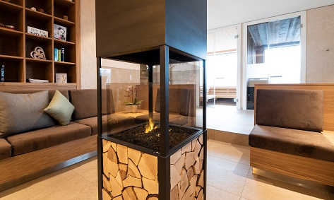 Moderner Loungebereich mit braunen Sofas, einem zentralen, verglasten Kamin, der mit gestapeltem Brennholz gefüllt ist, und Regalen mit Büchern und Dekoartikeln auf der linken Seite; helle, klare Atmosphäre.