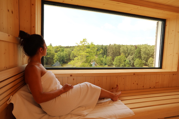 Eine in ein Handtuch gehüllte Frau entspannt sich auf einer Holzbank in einer Sauna und blickt durch ein großes Fenster auf eine malerische Aussicht mit grünen Bäumen und einem strahlend blauen Himmel. Im Peters HOtel & Spa in Jägersburg Saarland